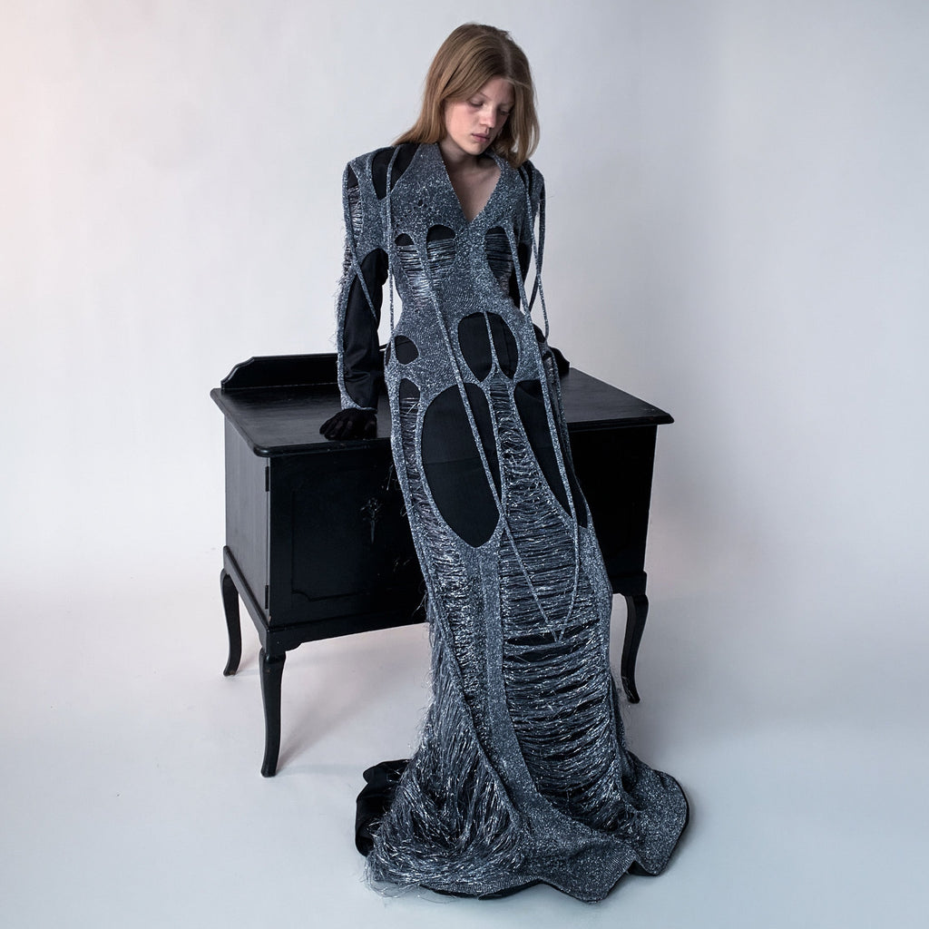 Woman wearing a long, dark gray dress layered with metallic knit standing next to a black desk.