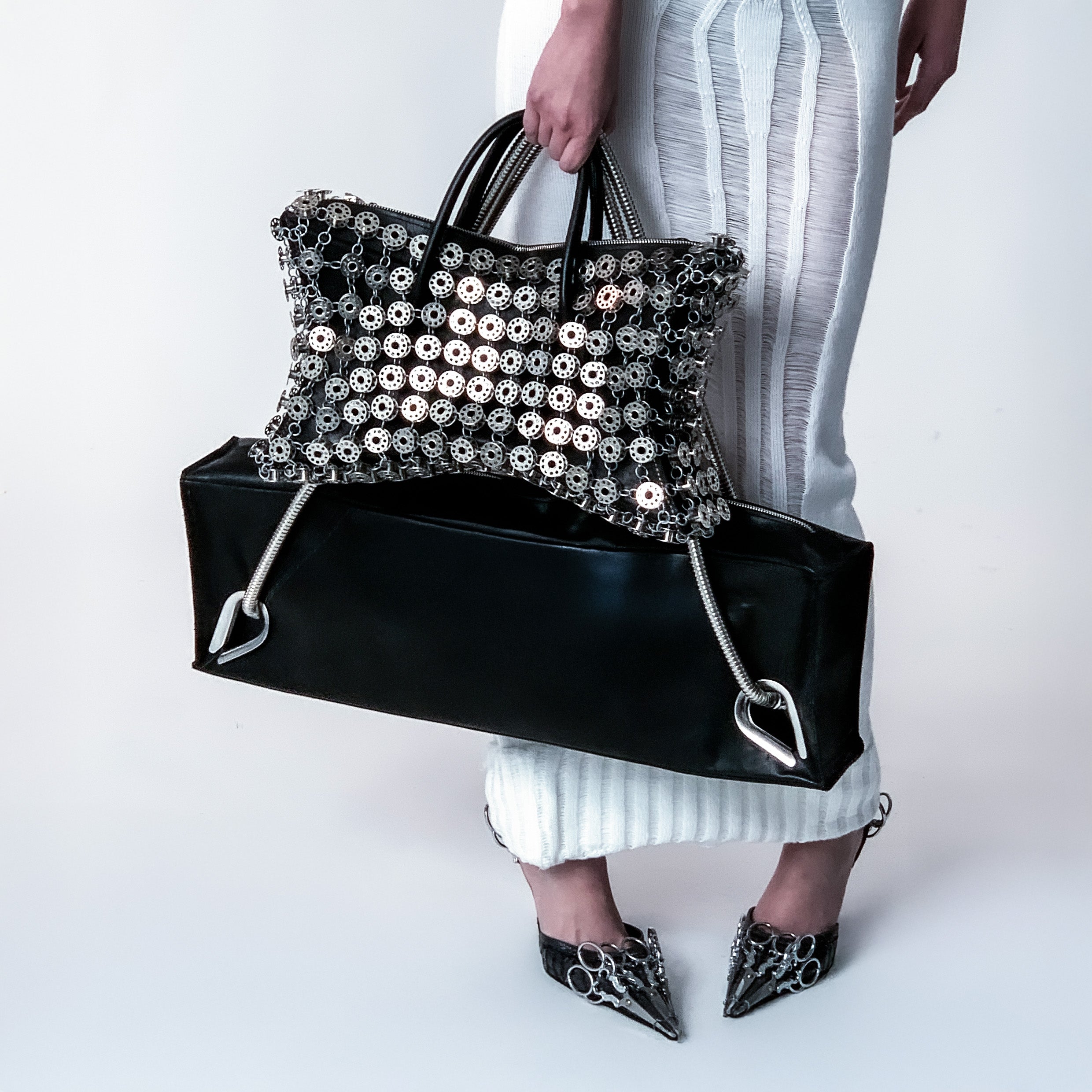 woman with a white knitted skirt holding a handbag layered with metal bobbins and a black leather bag with metal handles on light gray background 