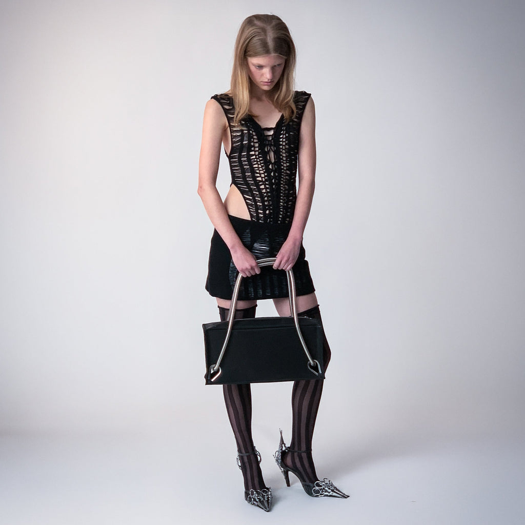 Person wearing a black knitted top, a black knitted skirt, upcycled black leather heals adorned with vintage metallic scissors holding a black leather bag with metallic handles on a light gray background