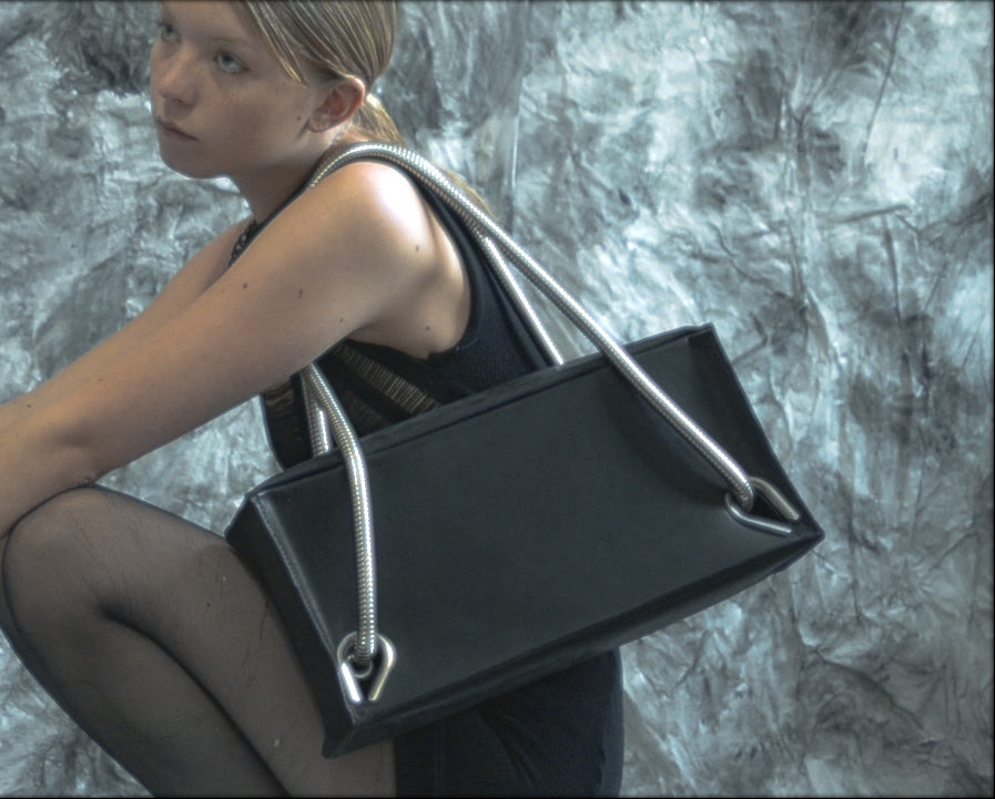 Person in a black knitted dress wearing a bllack leather handbag with silver handles and on a textured background 
